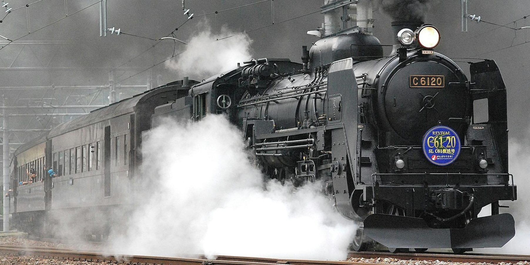 A powerful black steam locomotive, billowing thick black smoke and white steam, travels along railroad tracks under a network of overhead electric wires.