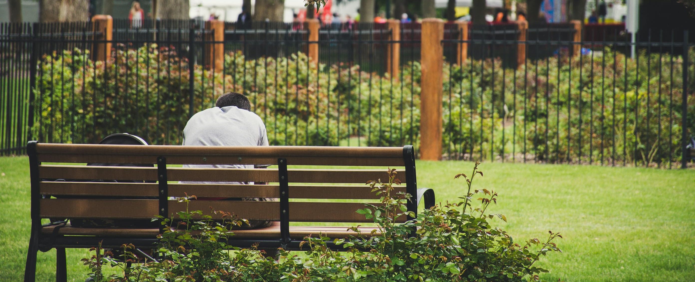 A person sits alone on a park bench, hunched over with their head down, surrounded by greenery and trees, with a blurred background of people and fences, evoking a sense of isolation and introspection.