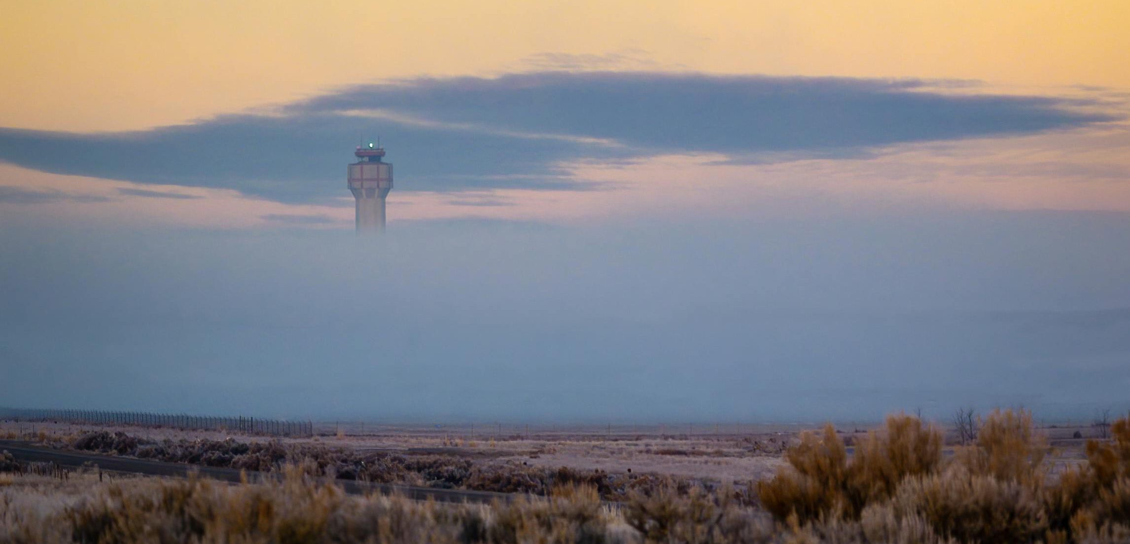 A control tower barely visible, rising through thick morning fog under a pastel sky of blue and gold hues.