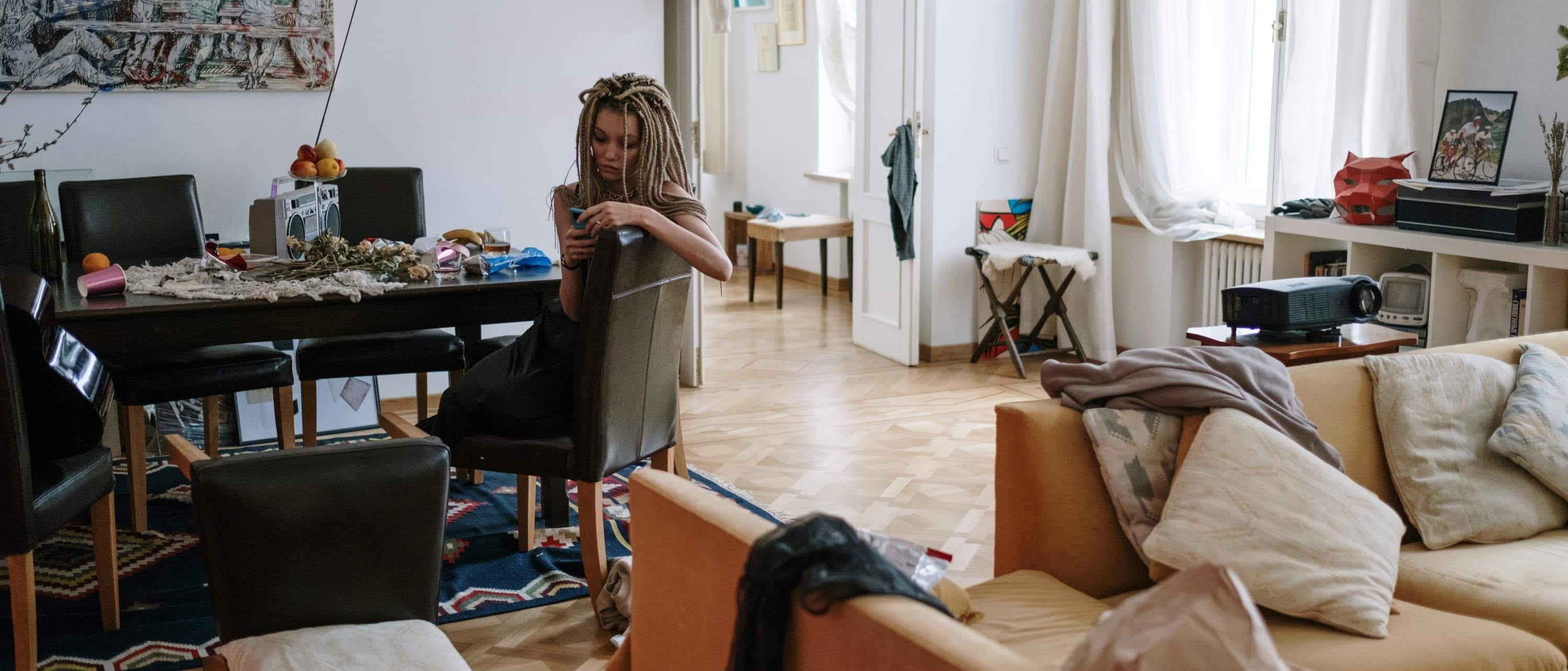 A young woman sits in a slightly messy living room, absorbed in her phone. Around her are scattered cushions, clothes draped over furniture, and a cluttered dining table, suggesting a sense of mental or emotional overwhelm.