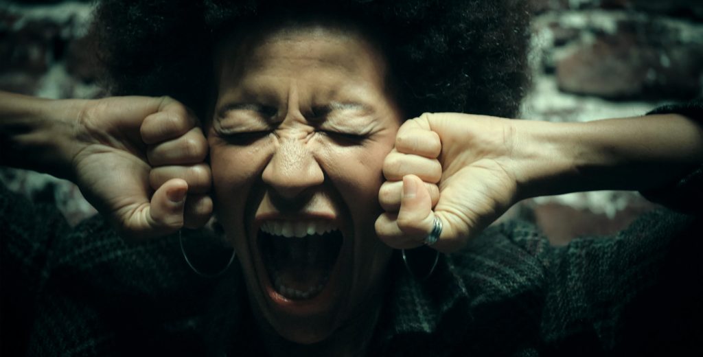 Close-up of a person screaming with fists clenched near their face, eyes shut tightly, expressing intense emotional overwhelm.