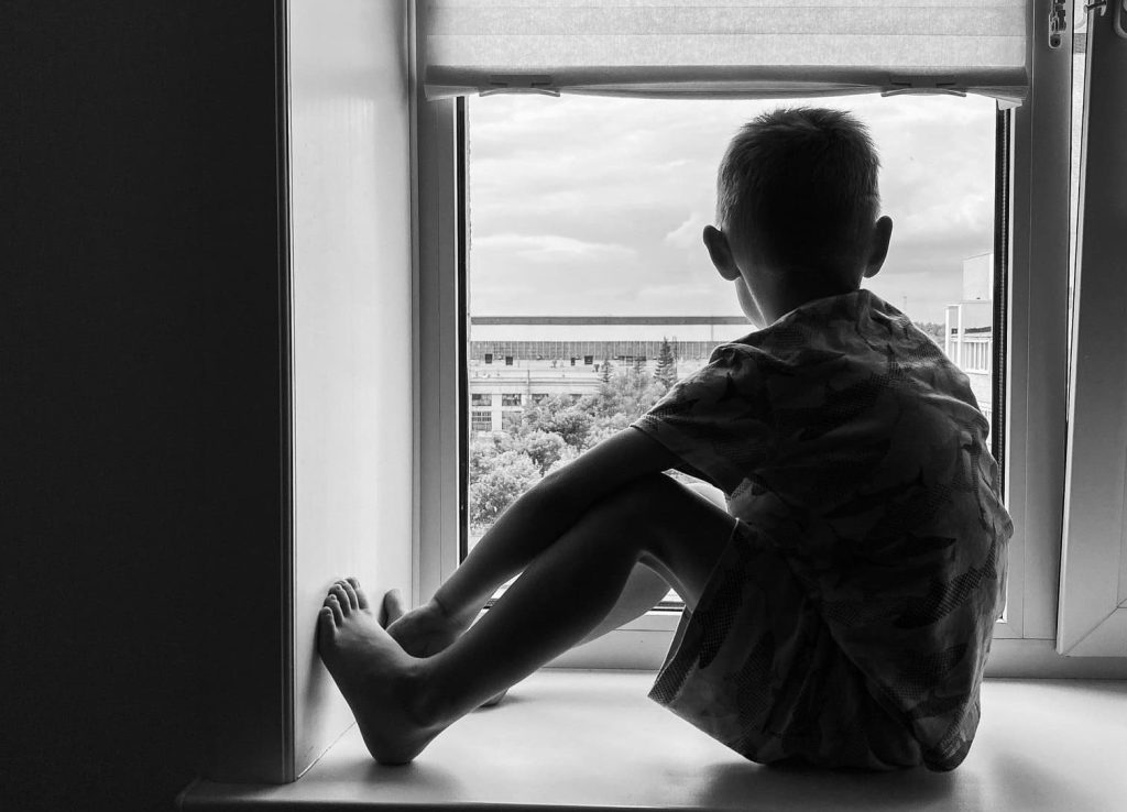 A young boy sits silently on a windowsill, gazing out at the distant cityscape. His posture is introspective, his small frame silhouetted against the light, capturing a quiet moment of solitude.