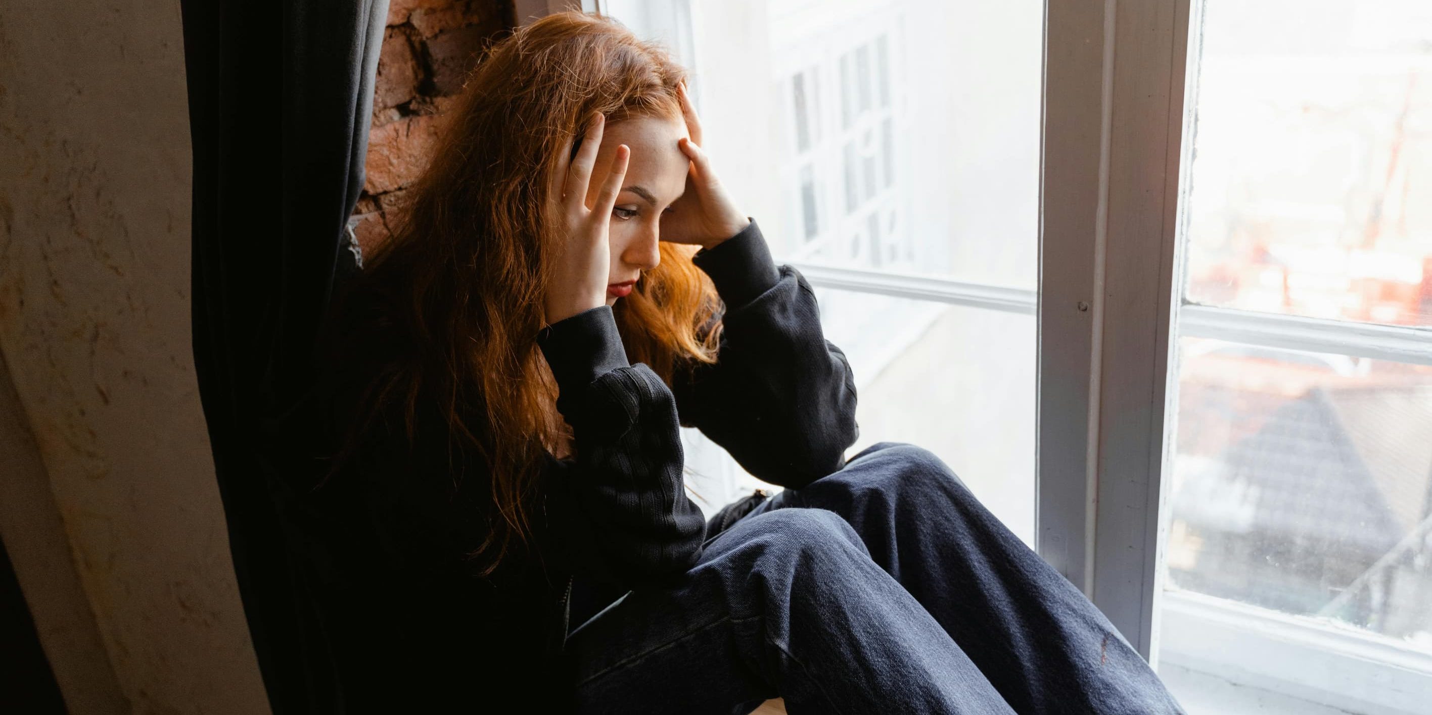 A young woman with red hair sits on a window ledge with her head in her hands, staring out the window with a tired and distressed expression.