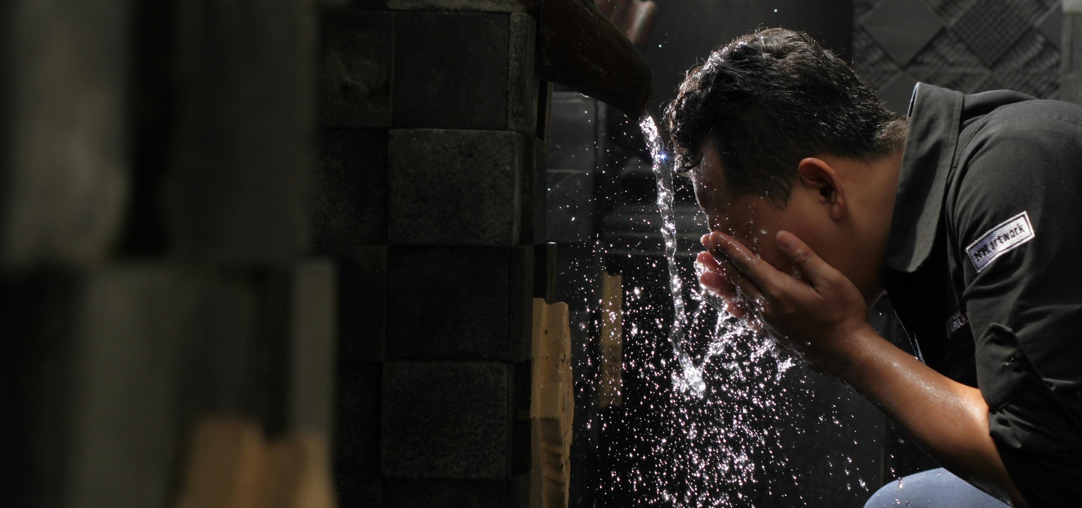 A man bends over and splashes water onto his face from a stone fountain, droplets captured mid-air as they scatter and reflect light.