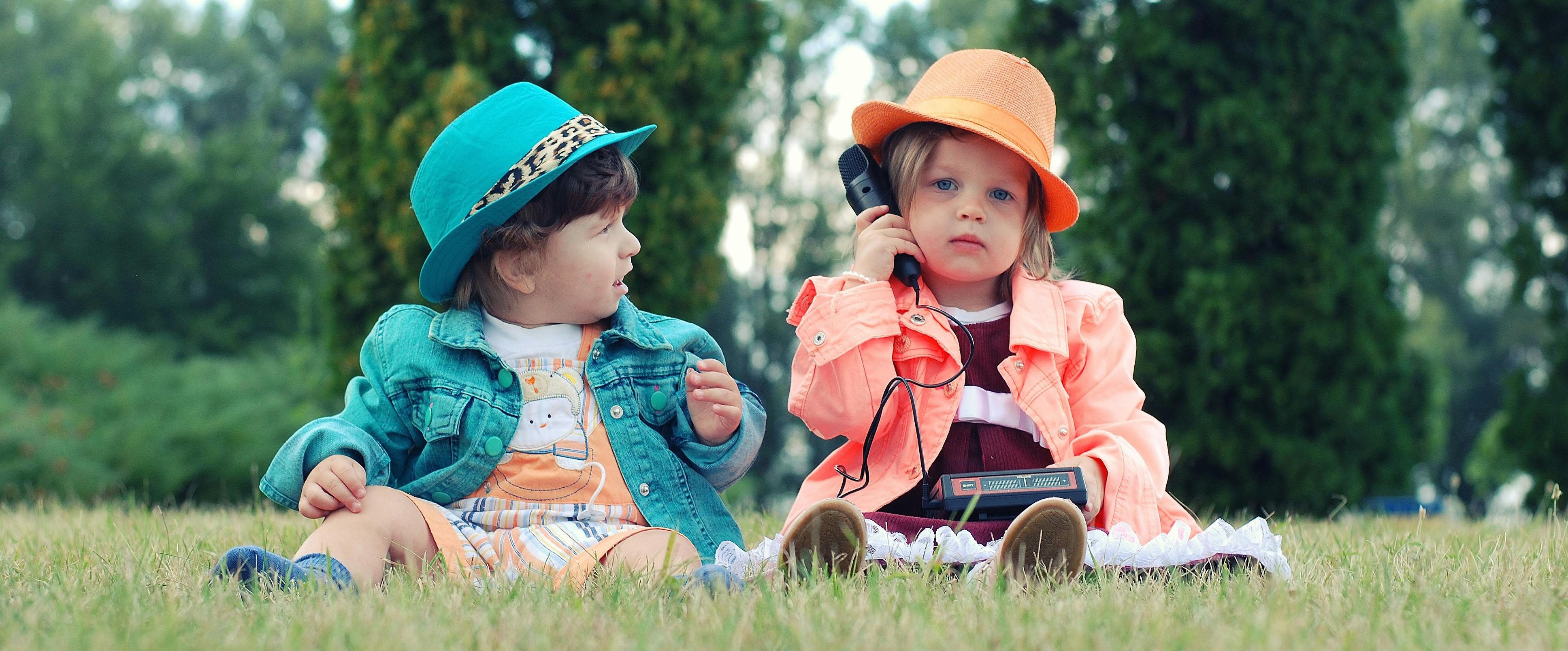 Two young children dressed in colorful outfits and stylish hats sit on the grass; one child holds a vintage phone while the other looks on curiously.