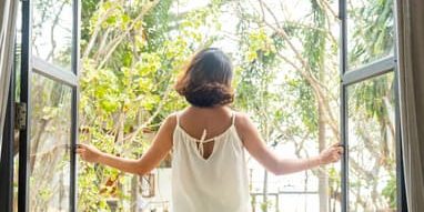 A woman in a white top stands at an open window, facing bright morning light and lush green trees outside.