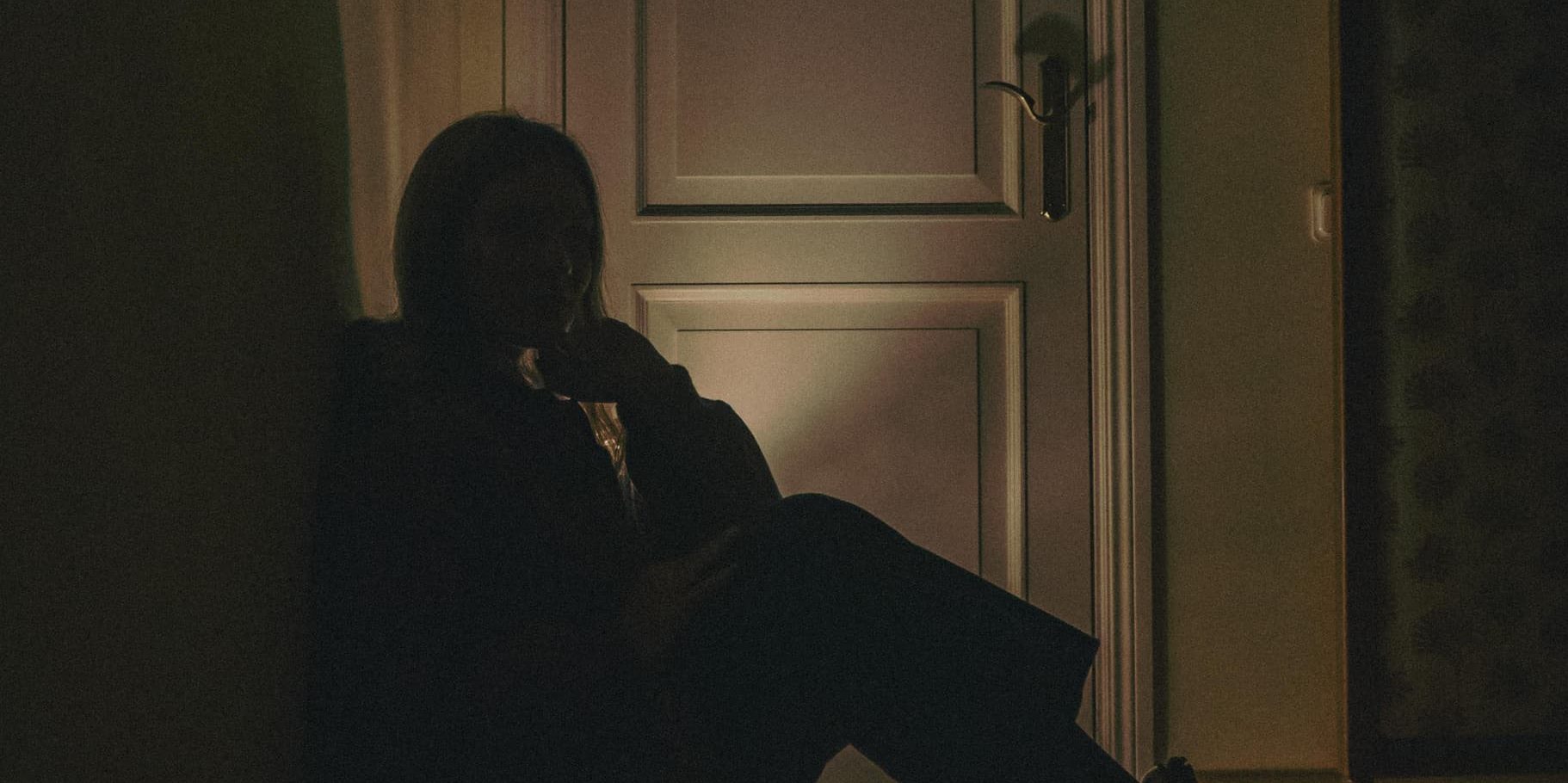 A person sits alone in a dimly lit room, their silhouette leaning against a wall near a closed door.