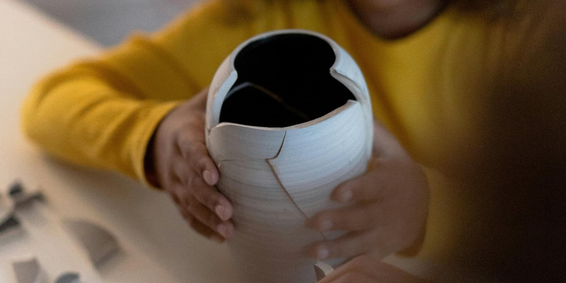A young child in a yellow shirt carefully holds a broken ceramic vase, attempting to fit the missing piece back together.