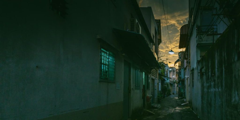 A dark alleyway between buildings, dimly lit by street lamps and the last light of dusk peeking through the clouds.