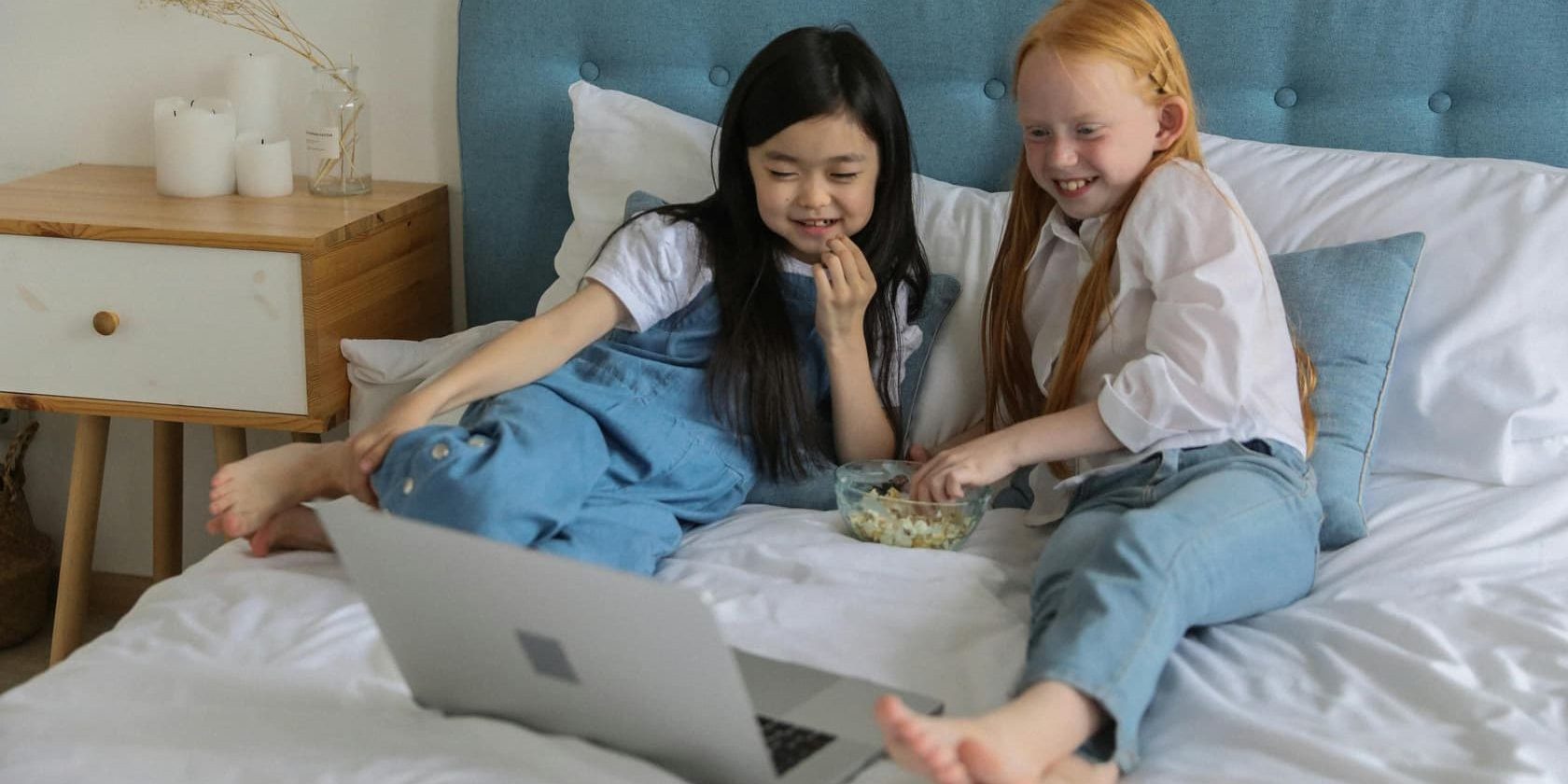 Two young girls sitting on a bed sharing popcorn and watching a laptop screen with joy and curiosity.