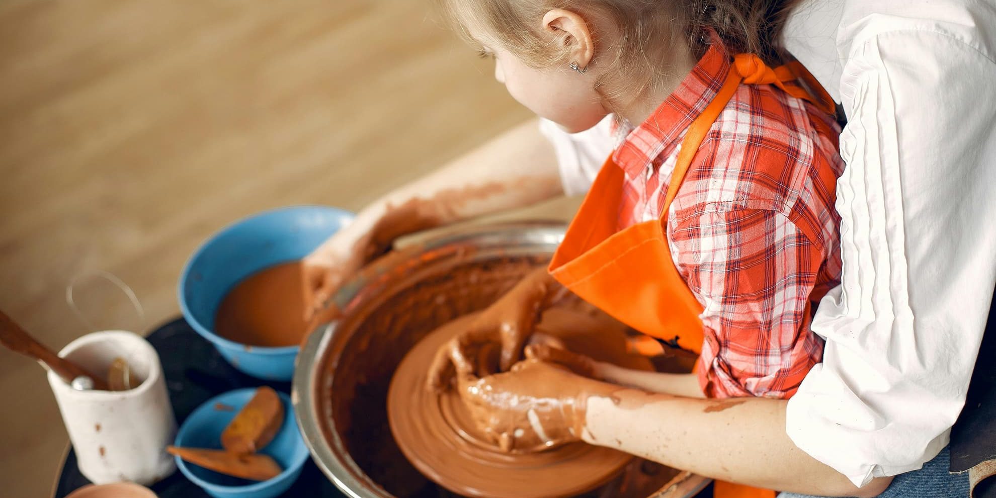 A parent helps a child shape clay on a pottery wheel, their hands layered in mud and motion.