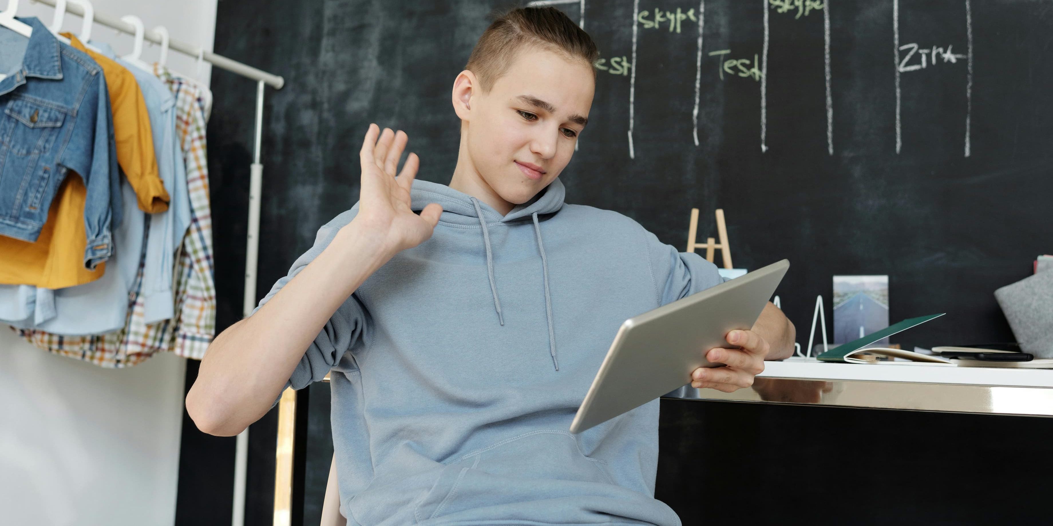 A teenager in a hoodie waves during a virtual class with a digital schedule written on a chalkboard behind him.