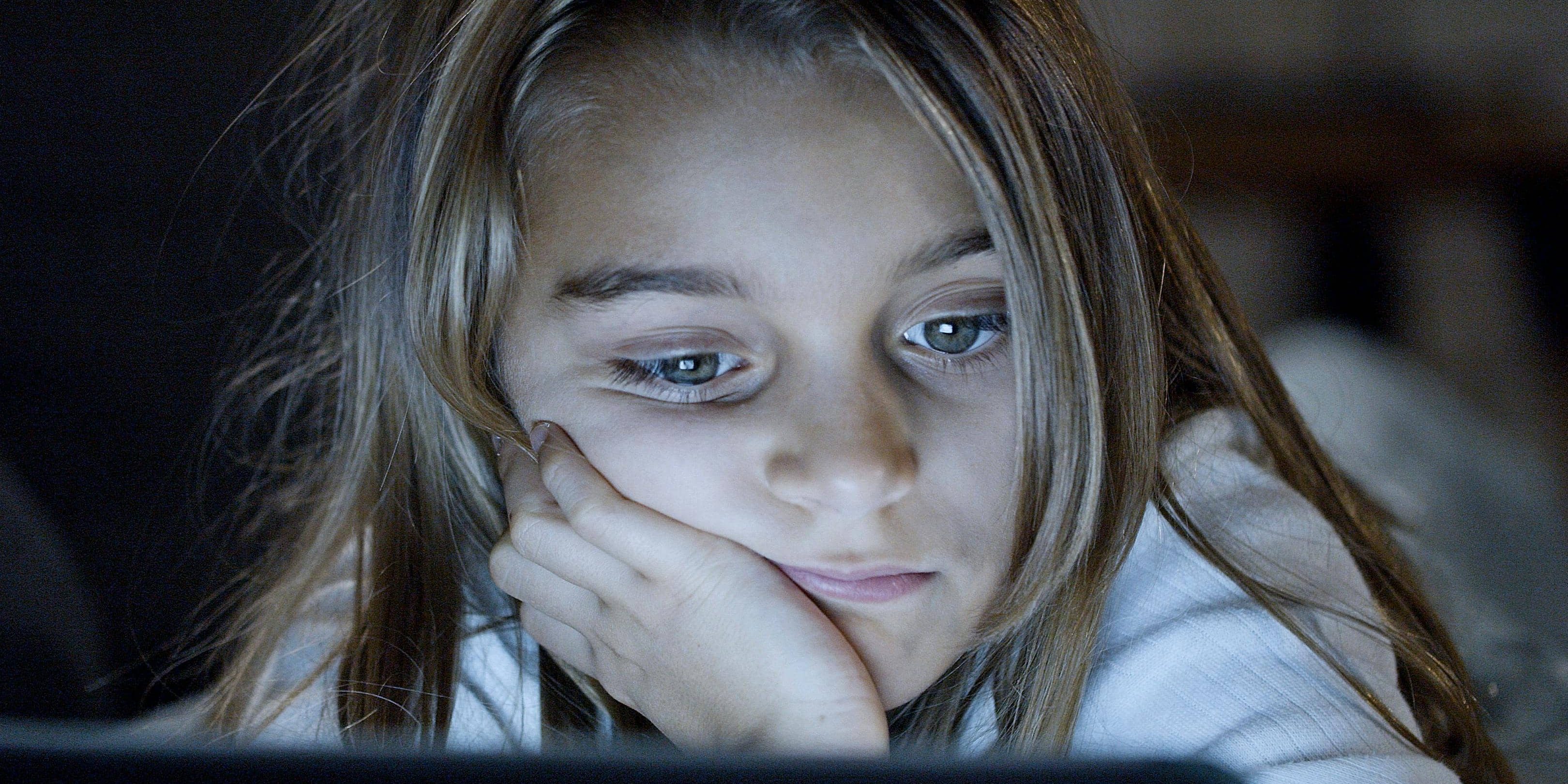 A young girl gazes at a screen in a dimly lit room, her expression distant and weary, with her head resting on her hand.