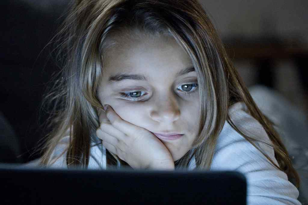 A young girl gazes at a screen in a dimly lit room, her expression distant and weary, with her head resting on her hand.