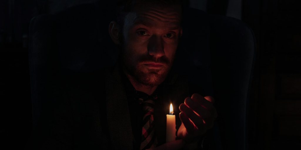 A solemn man sits in darkness, cupping a small candle with both hands. The gentle flame illuminates his face with soft, amber light, casting shadows that blend into the night around him.