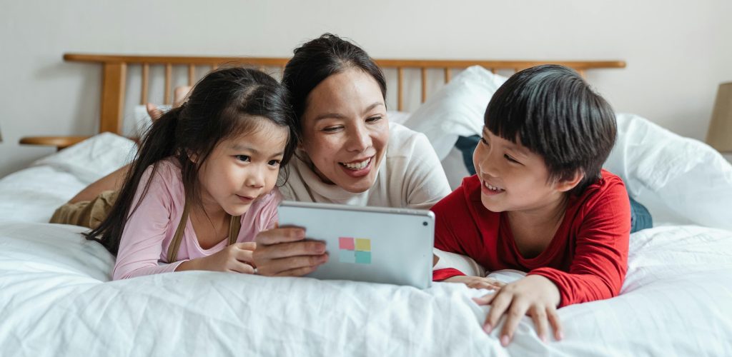 A smiling mother lying on a bed with her two children, enjoying a shared digital moment while watching a tablet together.