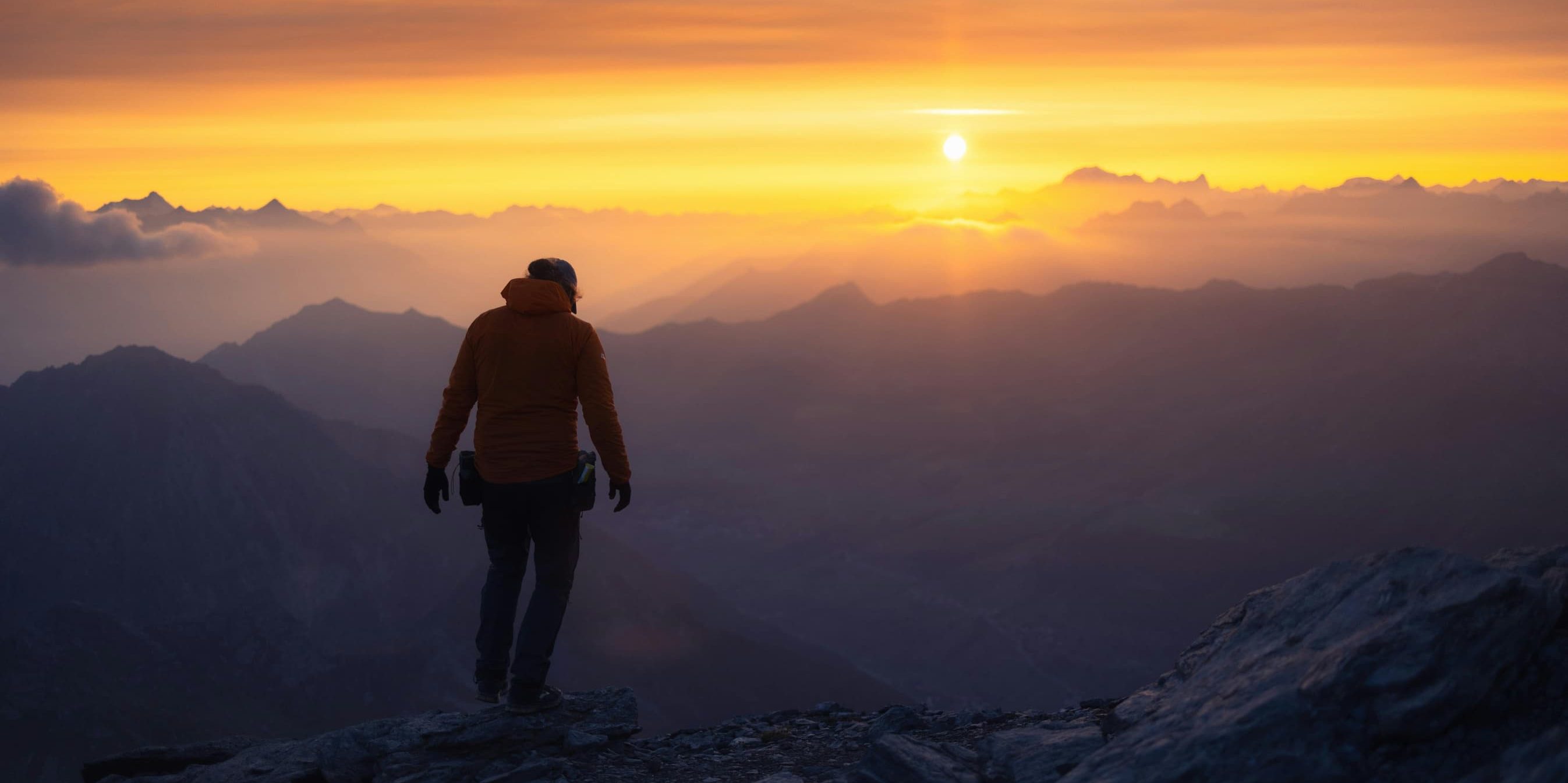 A lone hiker stands on a mountaintop at sunrise, gazing over misty valleys and distant peaks.