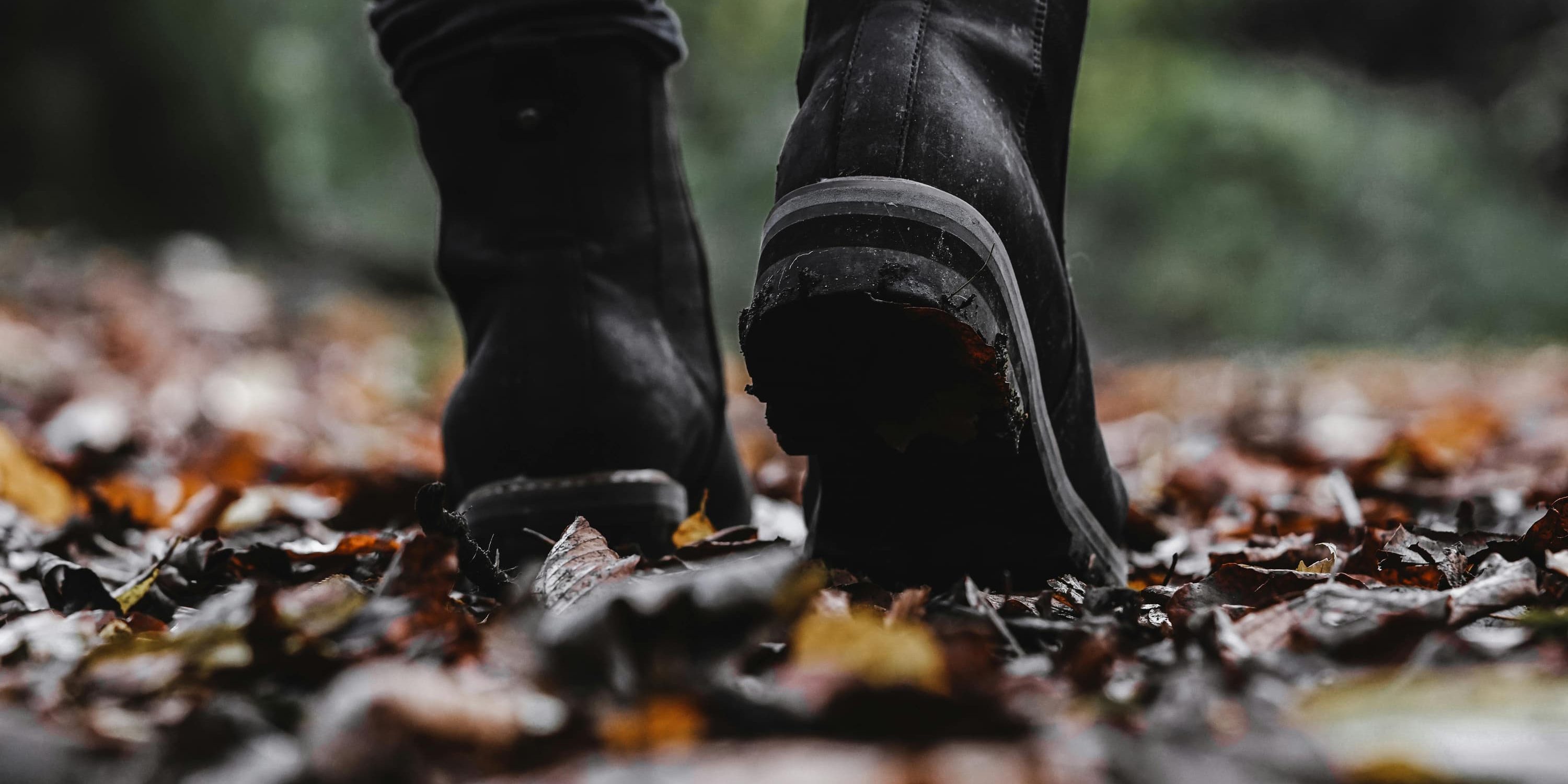 A close-up of black boots stepping through a forest path covered in wet autumn leaves, the focus on one sole mid-step, surrounded by a blur of earthy tones.