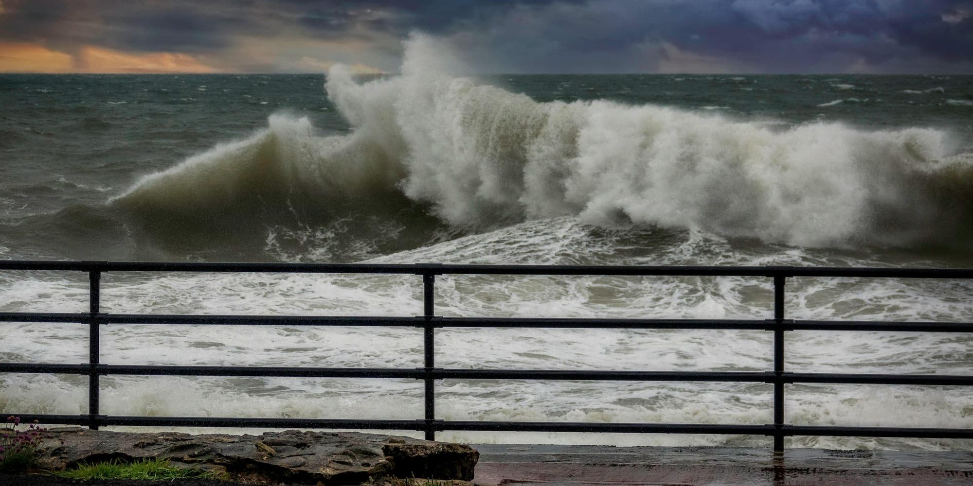 A stormy ocean wave crashing violently against a railing by the shore, with dark clouds looming above.