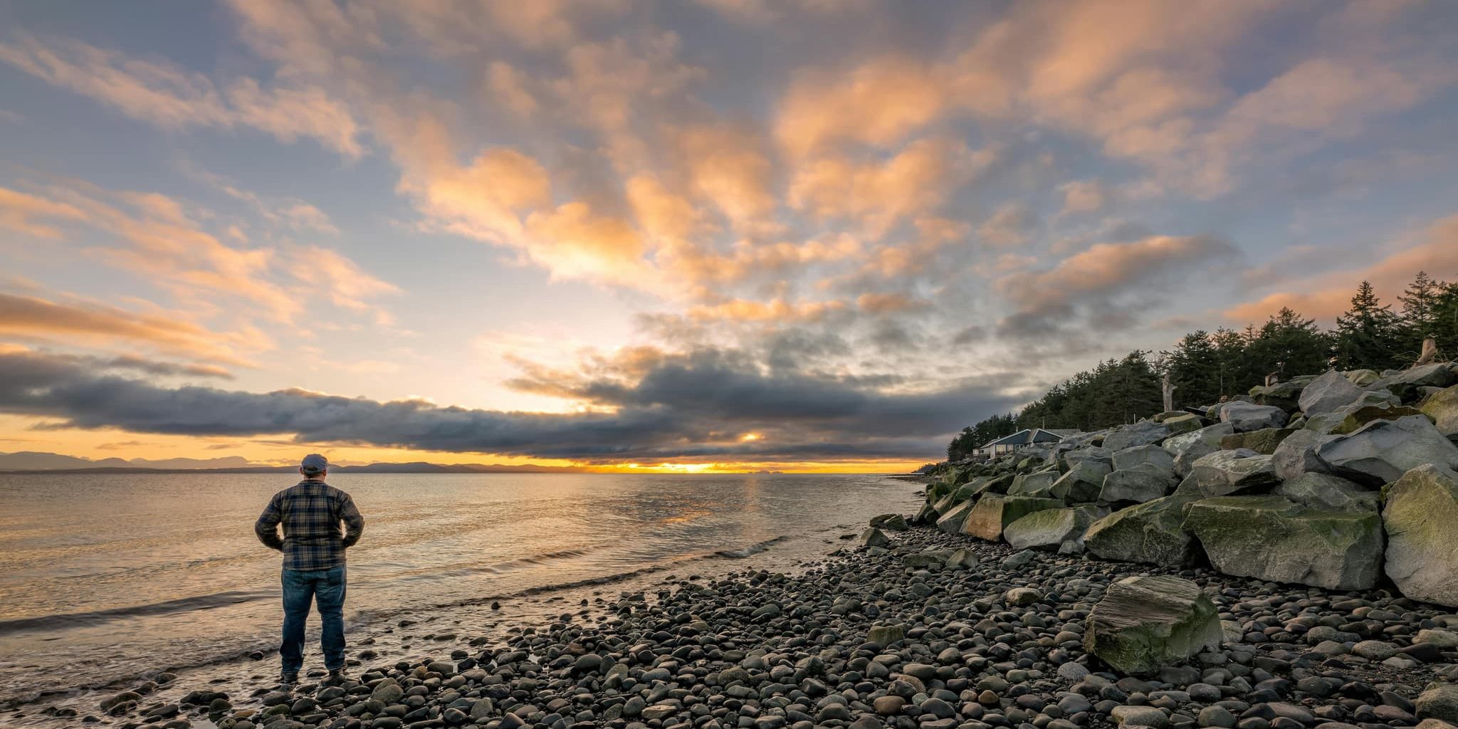 A solitary man stands on a rocky shore at sunrise, gazing toward the horizon where golden light pierces the clouds.