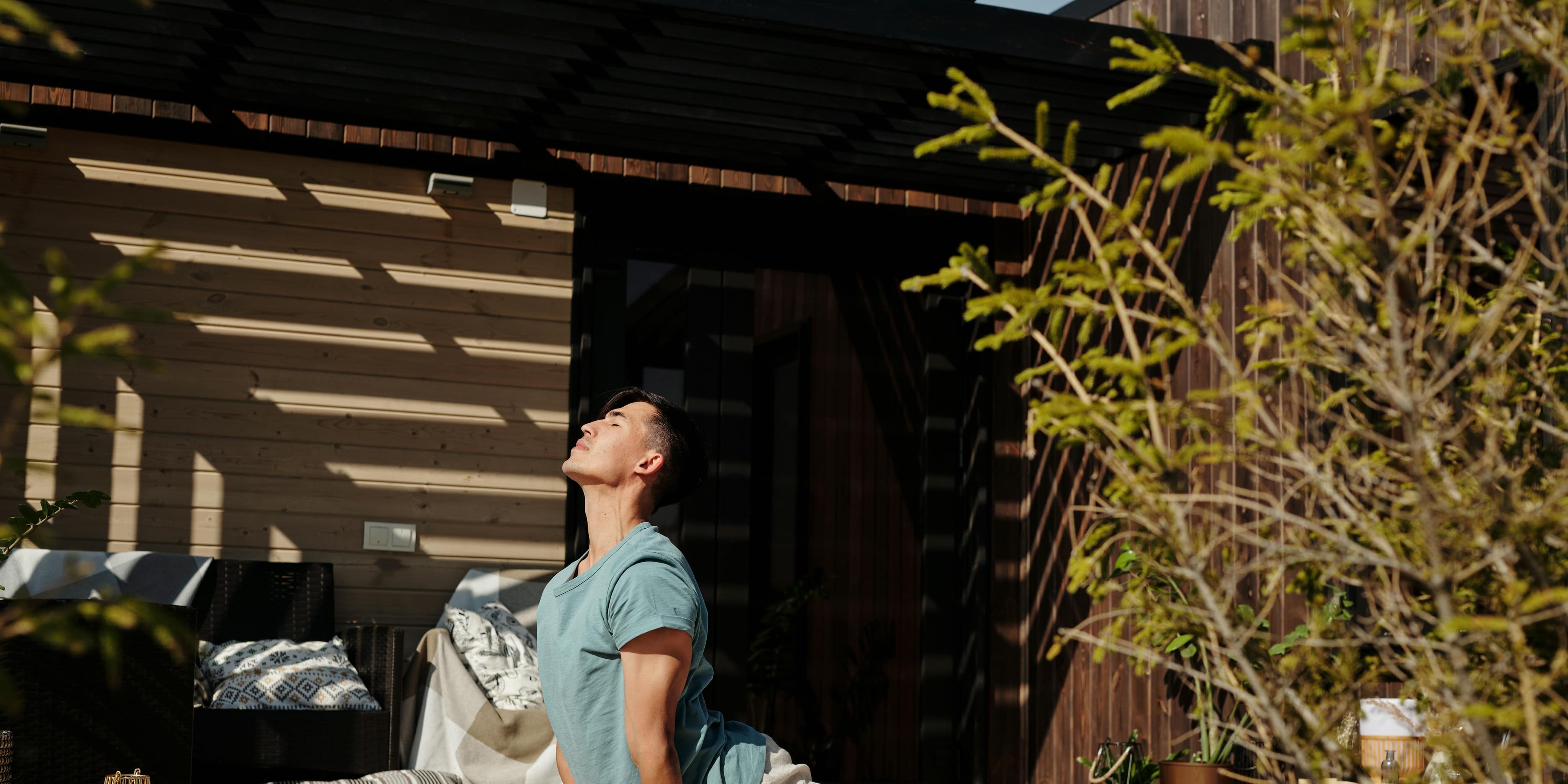 A man practices yoga in upward-facing dog pose on a wooden deck, basking in filtered sunlight beside a modern cabin.