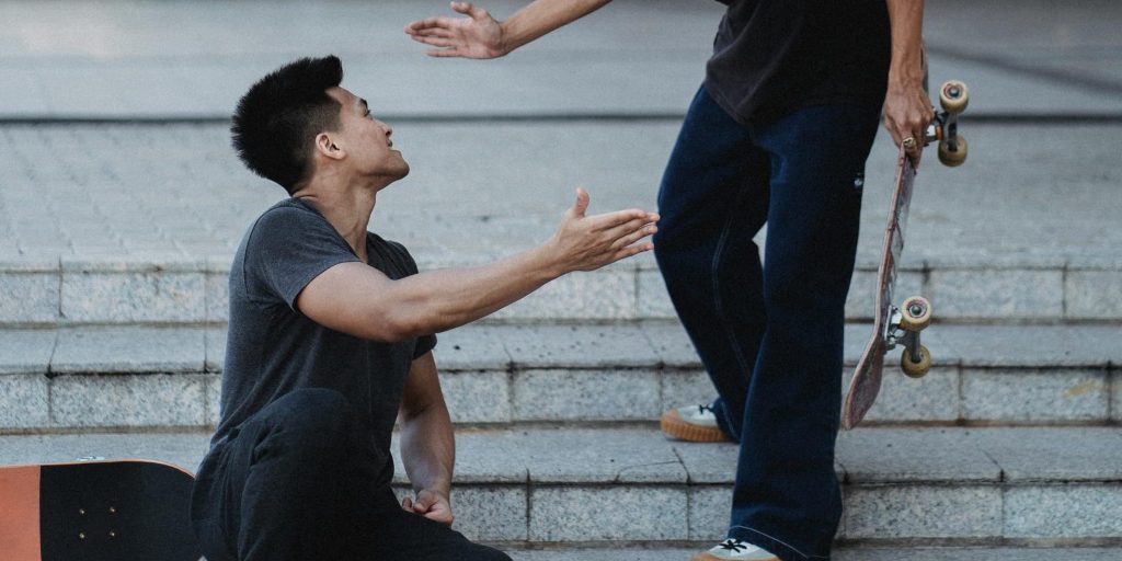 One skateboarder offers a hand to another who has fallen on stone steps, highlighting a gesture of support and camaraderie.