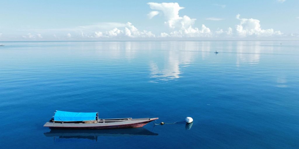 A single wooden boat with a blue canopy floats quietly on a vast, calm sea under a clear blue sky.