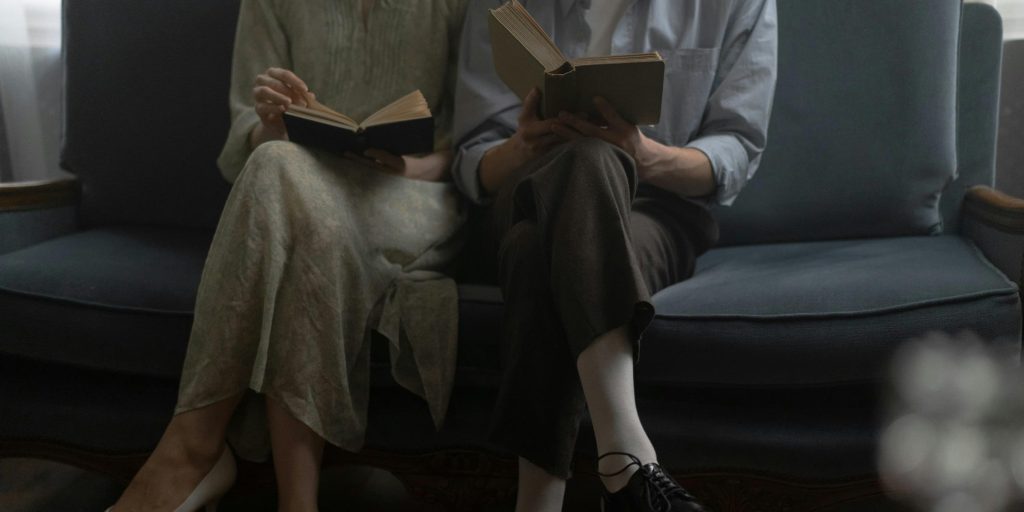 Two people sitting closely on a vintage sofa, each quietly reading a book, their legs slightly angled toward each other in silent companionship.
