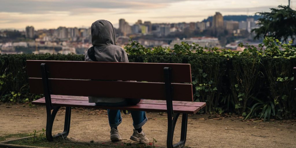 A solitary person in a grey hoodie sits on a park bench, gazing at a distant city skyline under a dusky sky.