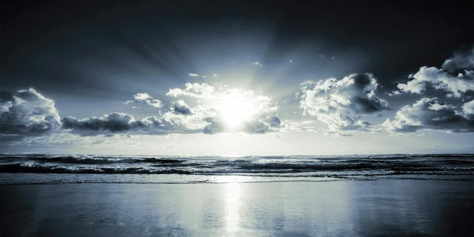 Sunlight breaking through dark clouds over a quiet ocean shoreline.