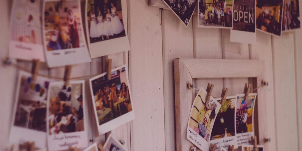A soft-focus image of polaroid-style photographs pinned with mini clothespins to string across a rustic white wooden wall; the photos capture joyful moments, people, and memories in warm tones.