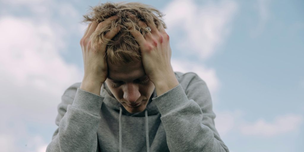 A young man in a hoodie clutching his head in distress against a cloudy sky.