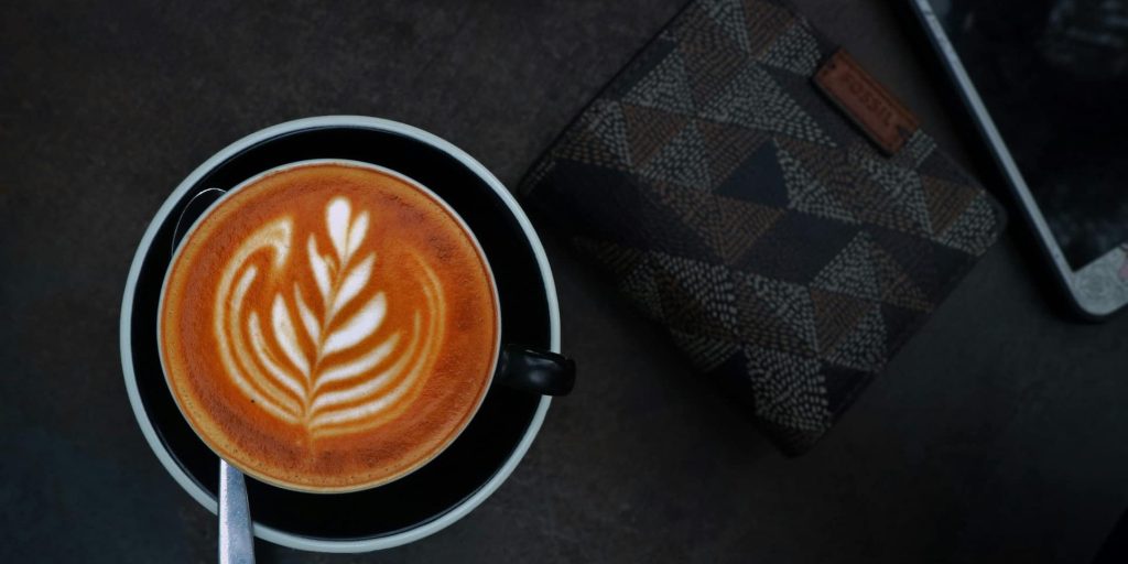 A top-down view of a cappuccino with elegant leaf latte art in a black cup, placed beside a patterned wallet and smartphone on a dark table.