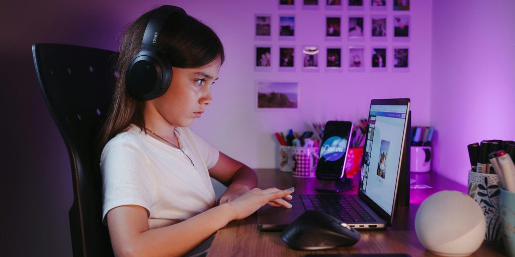 A young girl wearing large black headphones sits at a wooden desk, focused on her laptop screen in a softly lit purple room.