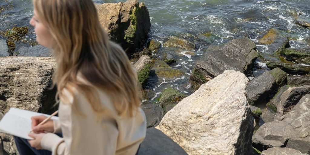 A person sitting on rocks near the water’s edge, journaling while waves crash gently against the shore.