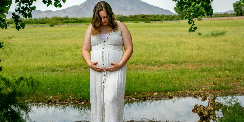 Pregnant woman in a white dress standing in a green field near a small pool of water, gently holding her belly under a tree arch with mountains in the background.