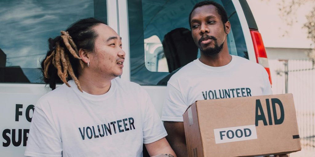 Two volunteers carrying aid and food boxes, standing together beside a van, ready to distribute supplies.