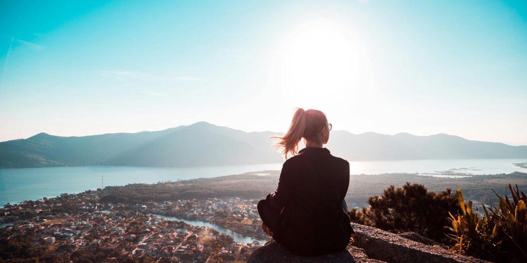 A woman sits on a rock overlooking a valley town, with mountains and a shining lake under the rising sun.