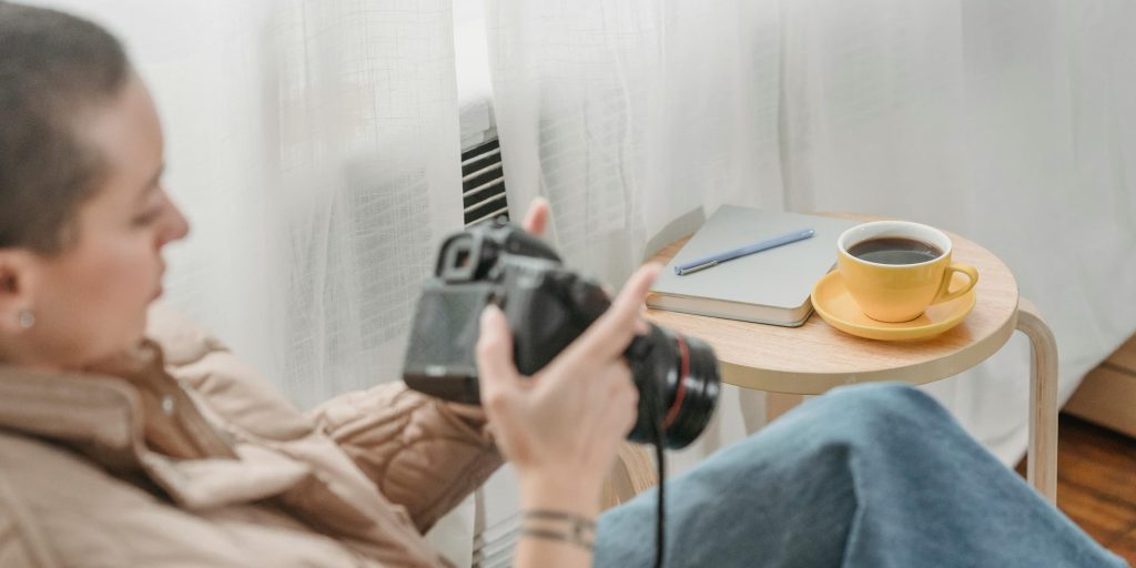 A woman holds a camera while sitting beside a table with coffee and a notebook.