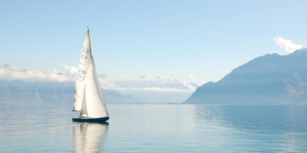 A lone sailboat gliding across still blue waters with distant mountains under a soft sky.