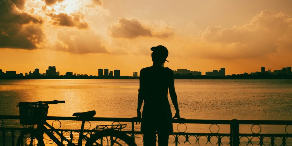 A person leans on a railing beside a bicycle, gazing at a golden sunset over a calm lake.
