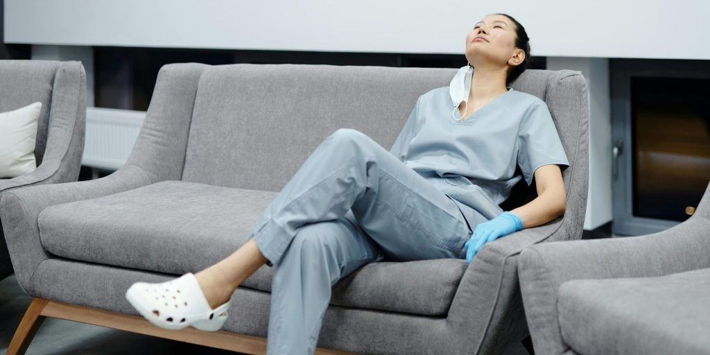 A tired healthcare worker in scrubs rests on a grey couch, leaning back with eyes closed, her mask lowered — a quiet moment of exhaustion and reflection after a long shift.