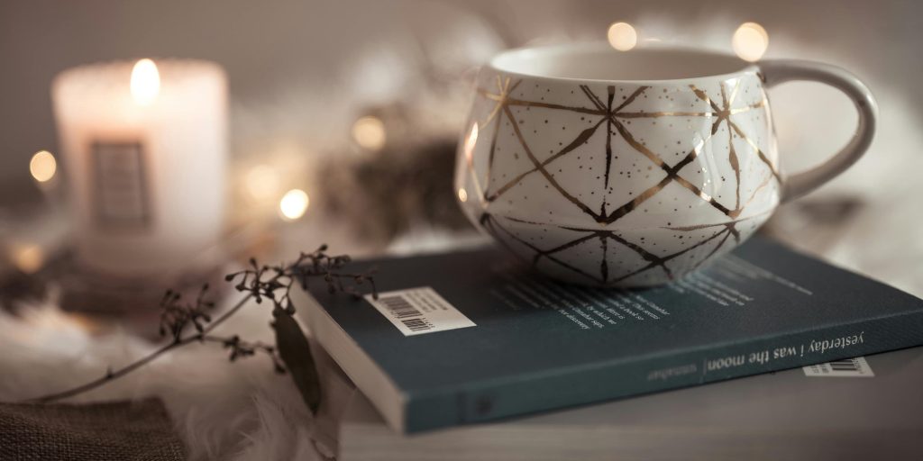 A cozy ceramic mug with golden geometric patterns rests on a closed book beside a softly glowing candle, surrounded by warm fairy lights.
