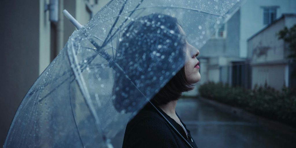 A woman stands beneath a clear umbrella in soft rain, her gaze lifted toward the sky as raindrops blur the city behind her.