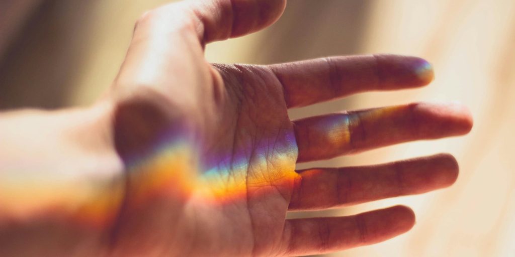 An open human hand bathed in sunlight, with a soft rainbow reflection cast across the skin.