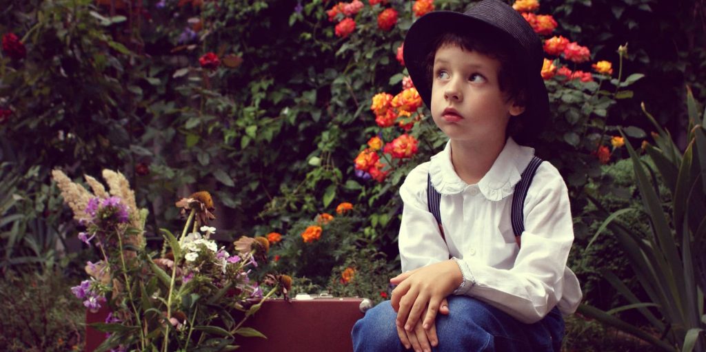 A young child in a white shirt and hat sits quietly among blooming flowers, gazing upward with a thoughtful expression.