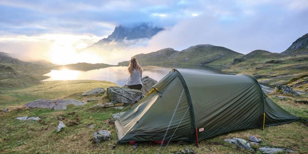 A lone camper sits beside a green tent overlooking a tranquil mountain lake at sunrise, surrounded by mist and soft golden light.