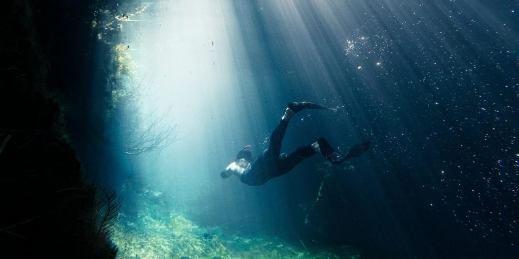 A lone diver descends into deep blue waters, surrounded by rays of sunlight streaming from above, creating a tranquil underwater glow.
