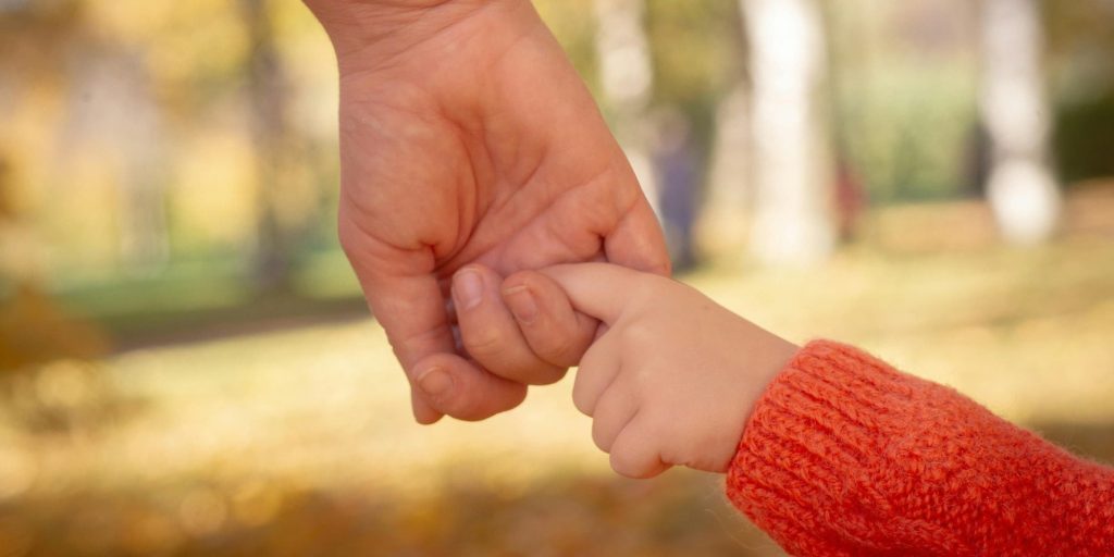 A close-up of an adult’s hand gently holding a child’s finger in a soft autumn light.